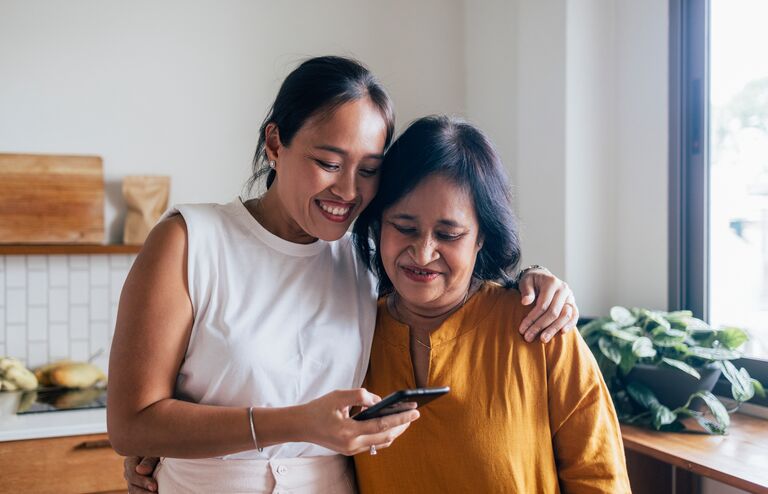 A smiling Asian female hugging her mother while they are reading something online using their black smartphone.