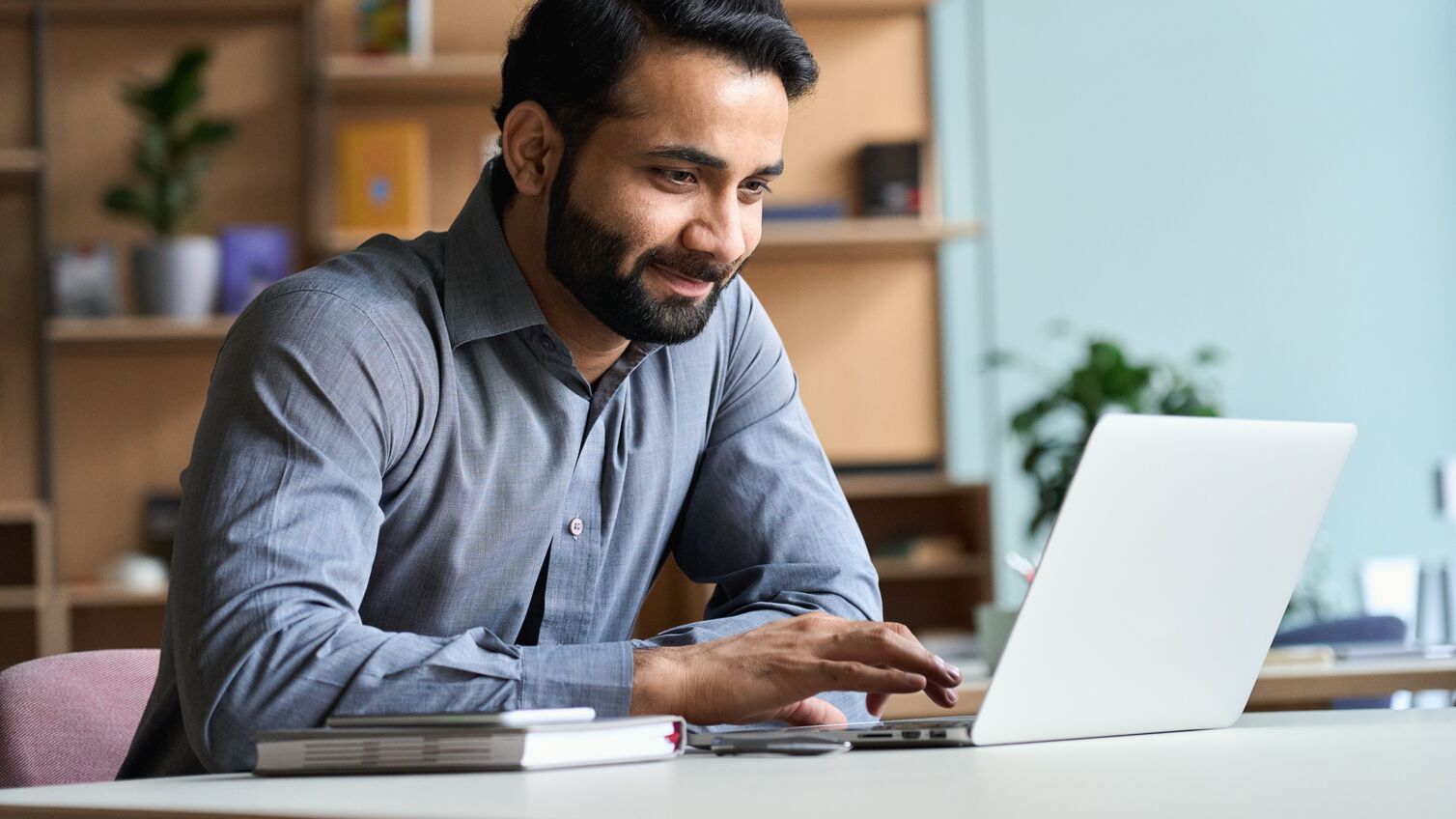 Smiling indian business man working on laptop at home office. Young indian student or remote teacher using computer remote studying, virtual training, watching online education webinar at home office.