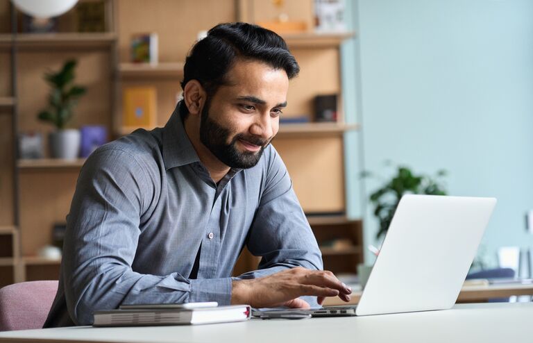 Smiling indian business man working on laptop at home office. Young indian student or remote teacher using computer remote studying, virtual training, watching online education webinar at home office.