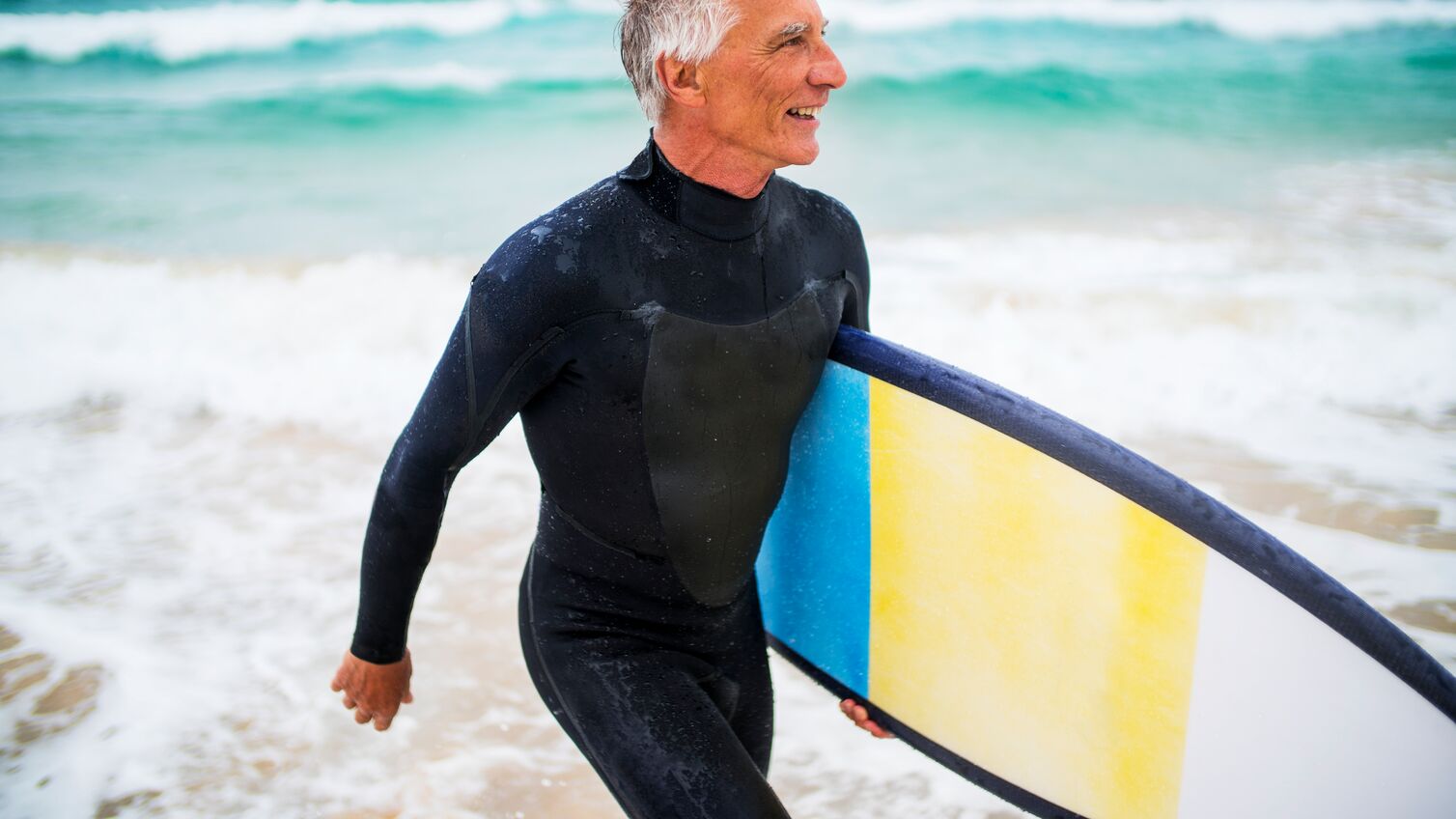 A senior man in wetsuit carrying surfboard on the beach. Bondi Beach, Sydney, Australia