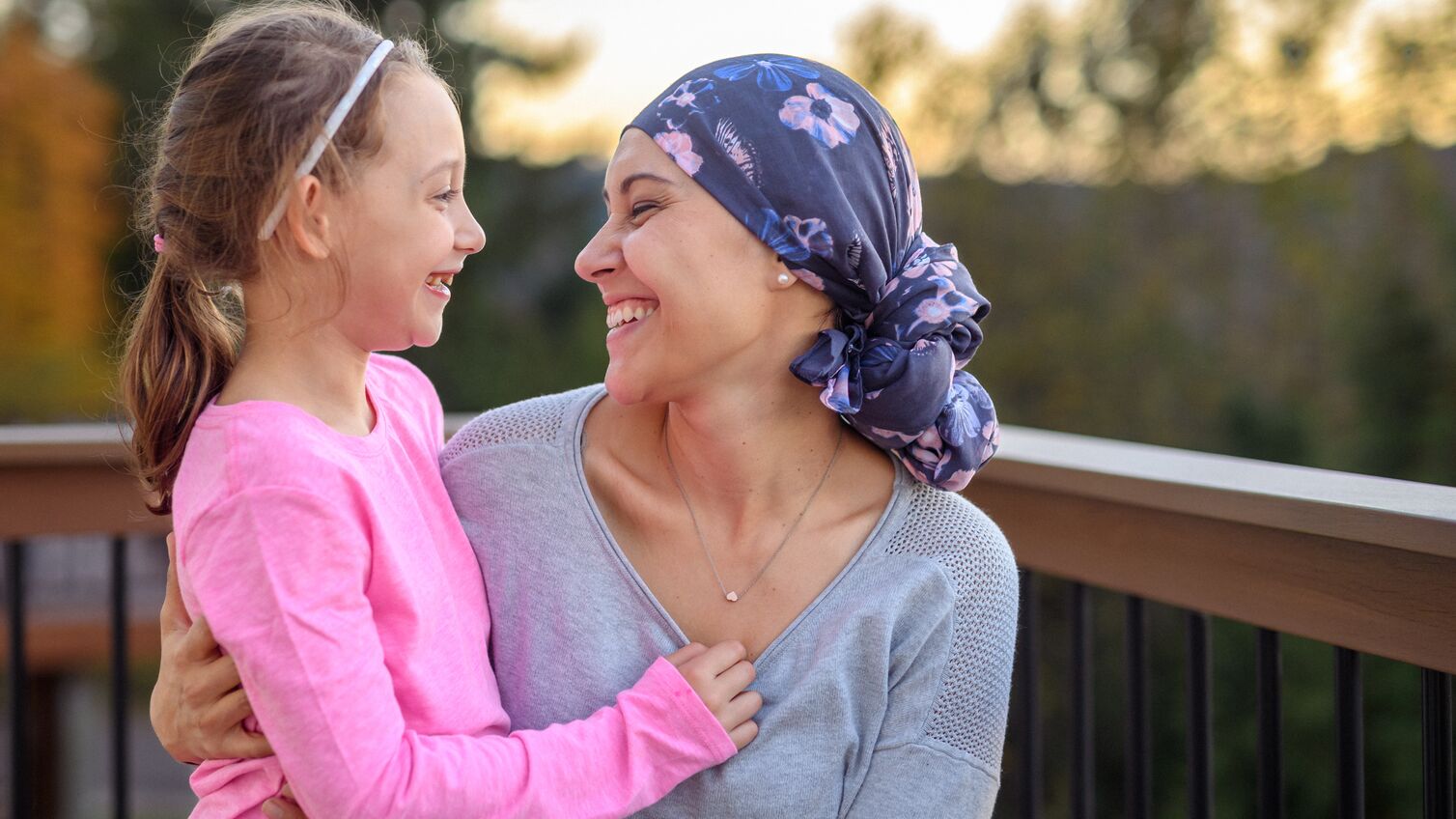 A cute elementary age girl and her mother look at each other and laugh joyfully after receiving the news that the woman's cancer treatment was successful and she is in remission. The family is spending time together outside on the back patio of their home on a warm and pretty evening.
