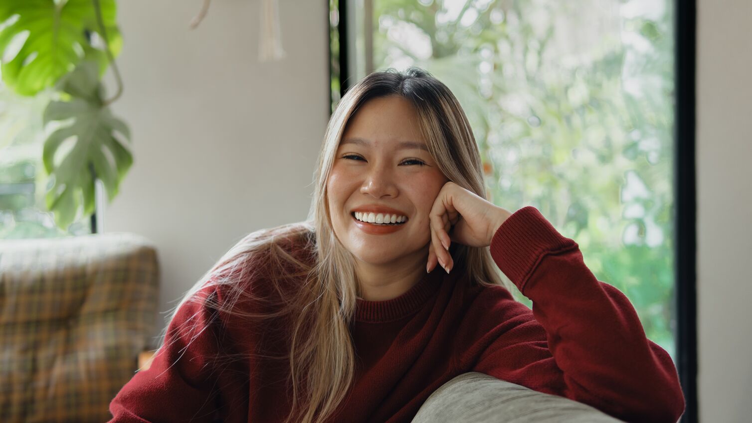 Happy young woman sitting in a cozy sofa, enjoying time with a smartphone. background with natural light and decorated with green plants, creating a warm and inviting atmosphere. Ideal for illustrating themes of social media, connectivity, and joyful moments in a relaxed setting.