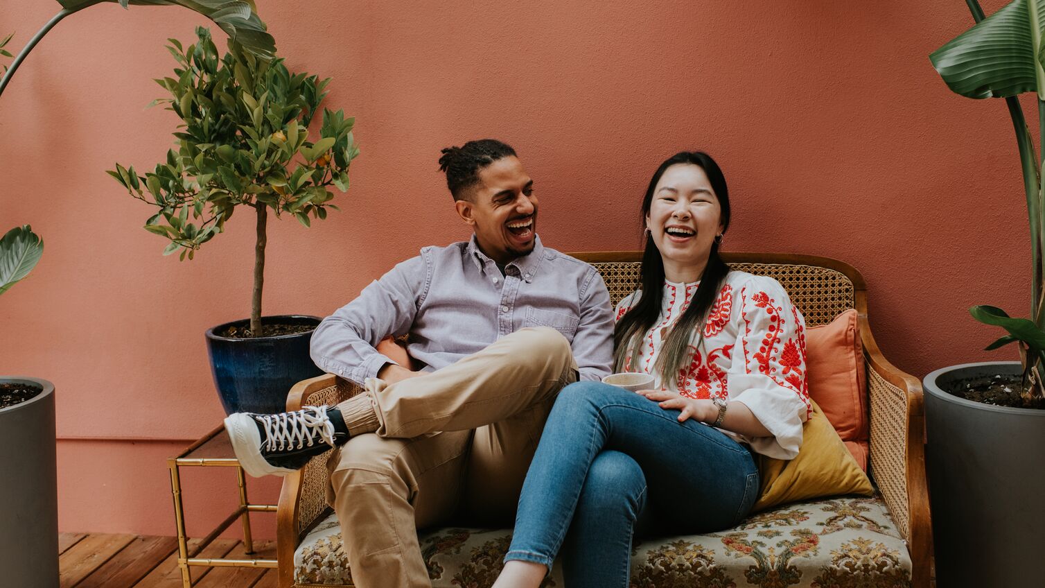 A black man and a Chinese woman sit side by side on a rattan sofa. They are comfortable together as they laugh. Space for copy.