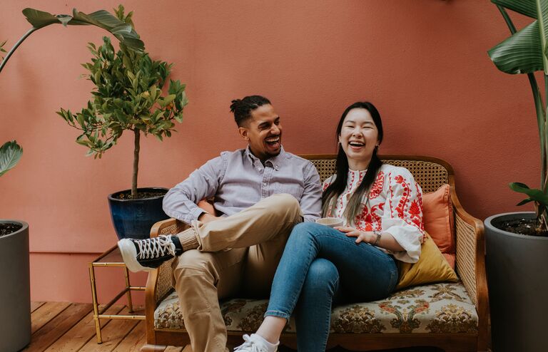 A black man and a Chinese woman sit side by side on a rattan sofa. They are comfortable together as they laugh. Space for copy.