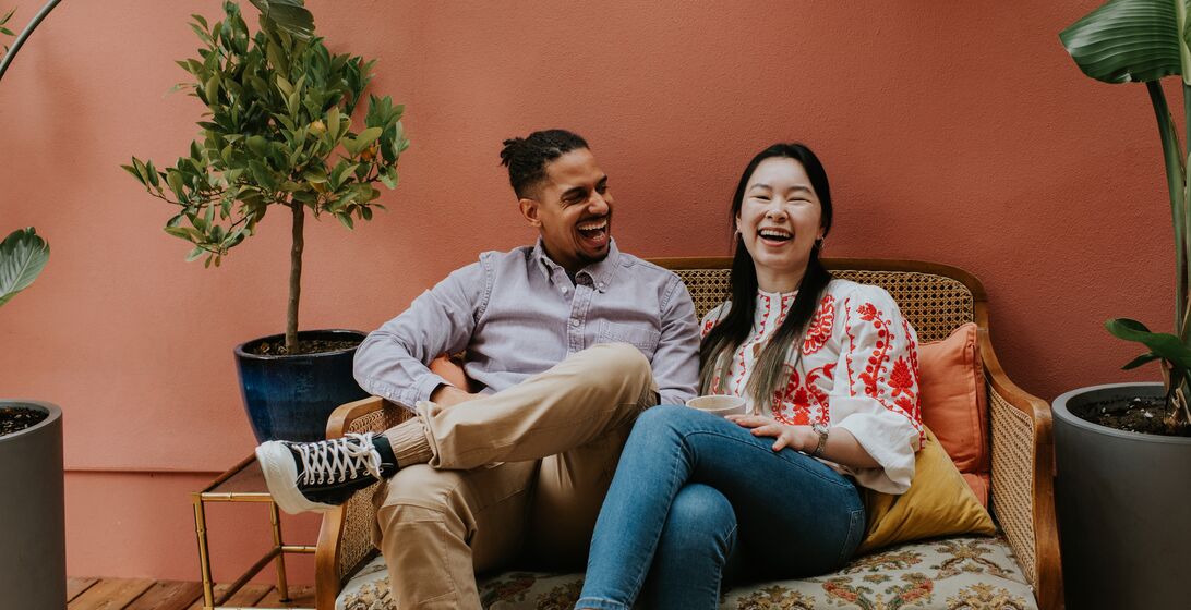 A black man and a Chinese woman sit side by side on a rattan sofa. They are comfortable together as they laugh. Space for copy.