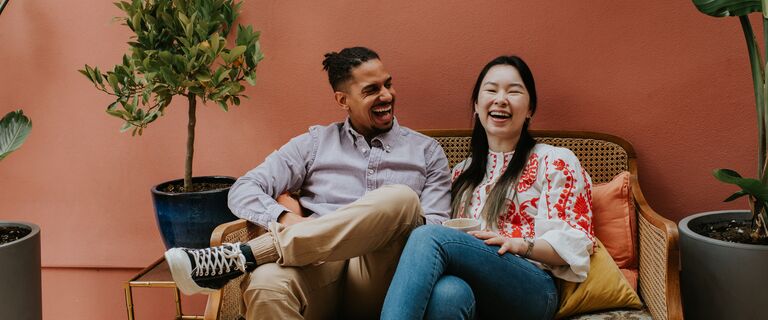 A black man and a Chinese woman sit side by side on a rattan sofa. They are comfortable together as they laugh. Space for copy.