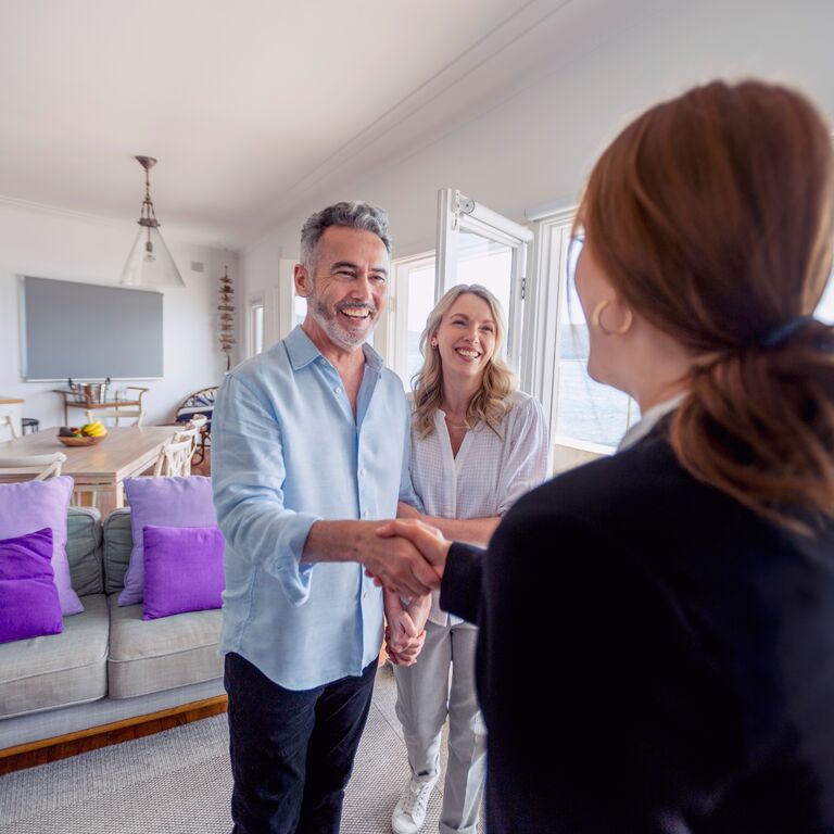 Real estate agent showing a mature couple a new house. The house is contemporary. All are happy and smiling. The couple are casually dressed and the agent is in a suit. The property is waterfront with the sea in the background.