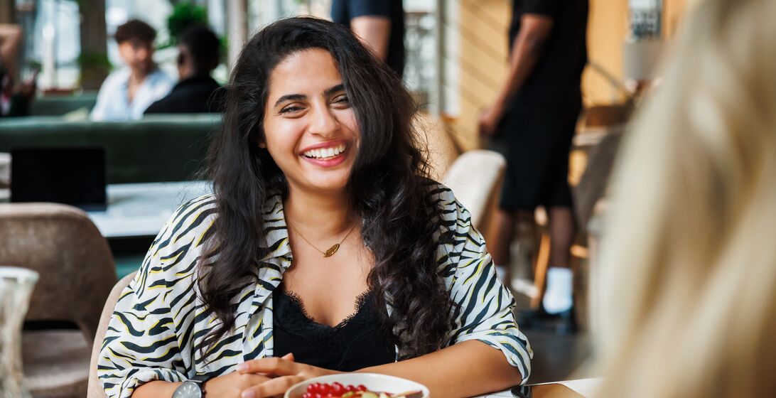A cheerful woman with a radiant smile enjoys a bowl of fresh fruit in a vibrant café setting, exemplifying a moment of urban leisure and healthy living.