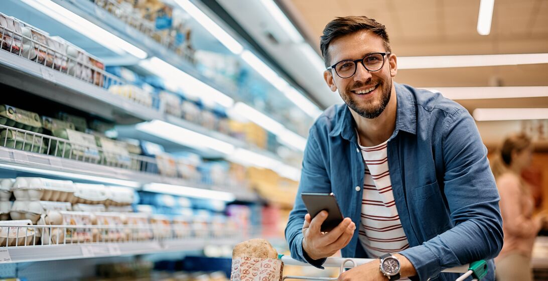 Happy man using smart phone while shopping in supermarket and looking at camera.