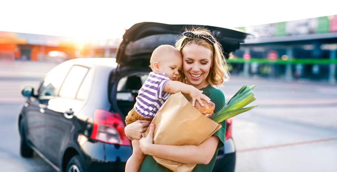 Beautiful young mother with her little baby son in front of a supermarket, holding paper shopping bag. Woman with a boy standing by the car.