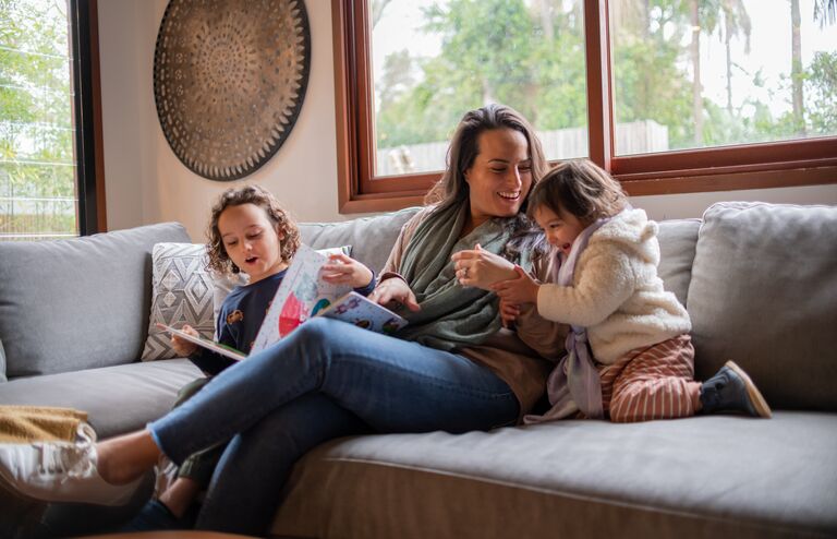 A mother laughs and plays with her two young children. They are snuggled up on the couch together and enjoying quality family time.