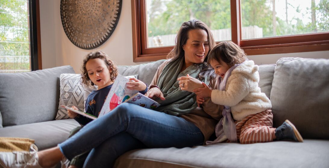 A mother laughs and plays with her two young children. They are snuggled up on the couch together and enjoying quality family time.