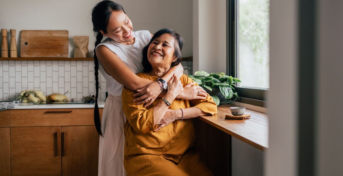 A smiling Asian woman enjoying drinking tea while her loving daughter is embracing her.