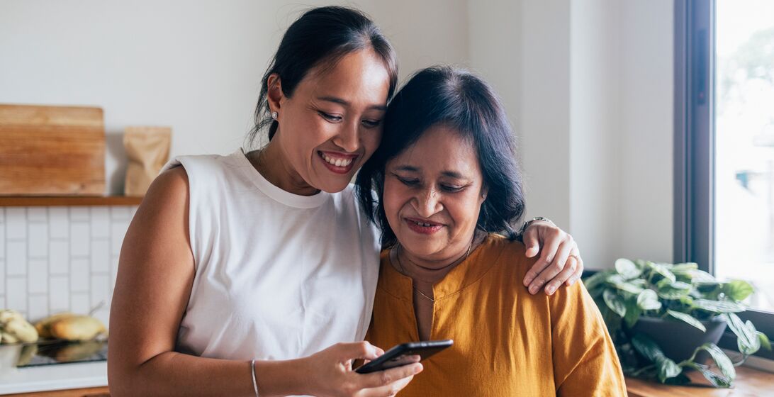 A smiling Asian female hugging her mother while they are reading something online using their black smartphone.