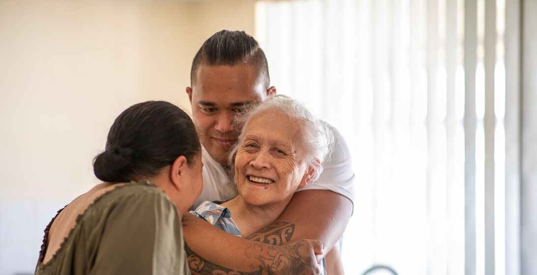Senior Pacific Islander woman and her mature daughter and grandson cuddling each other in their kitchen at home.