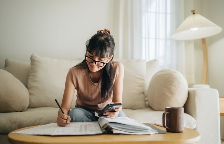 A woman sits at her living room with smartphone and financial reports doing her monthly budget.