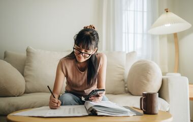 A woman sits at her living room with smartphone and financial reports doing her monthly budget.