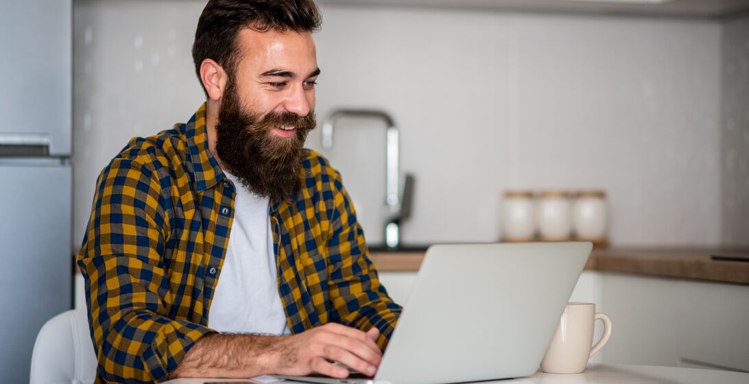 One young man is sitting in kitchen and typing on his laptop. He is freelancer and he works from home.