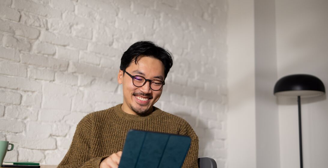 Young hip Chinese man sitting at his home office while enjoying his work using tablet