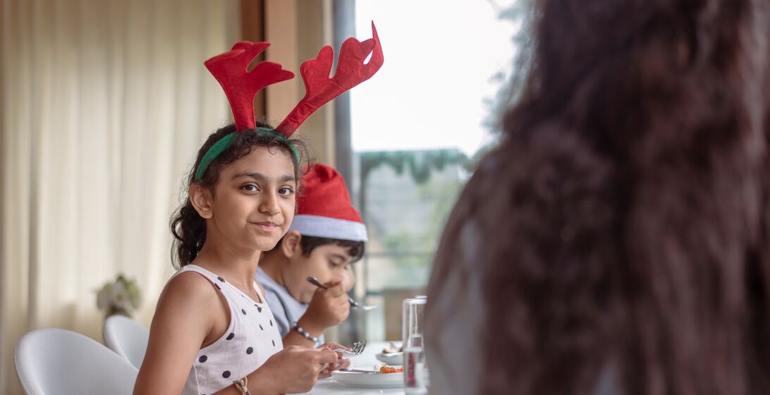 A young girl of Indian descent wearing a festive reindeer antler headband smiles directly at the camera while enjoying Christmas dinner with her family at home.