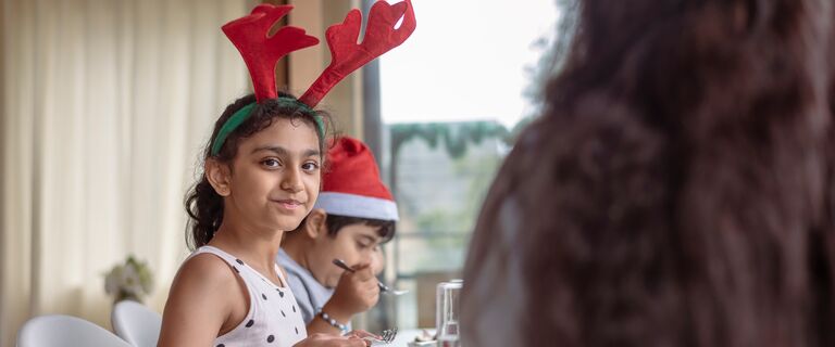 A young girl of Indian descent wearing a festive reindeer antler headband smiles directly at the camera while enjoying Christmas dinner with her family at home.