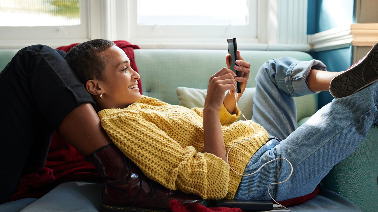 Smiling woman text messaging on smart phone while leaning on friend's leg in living room