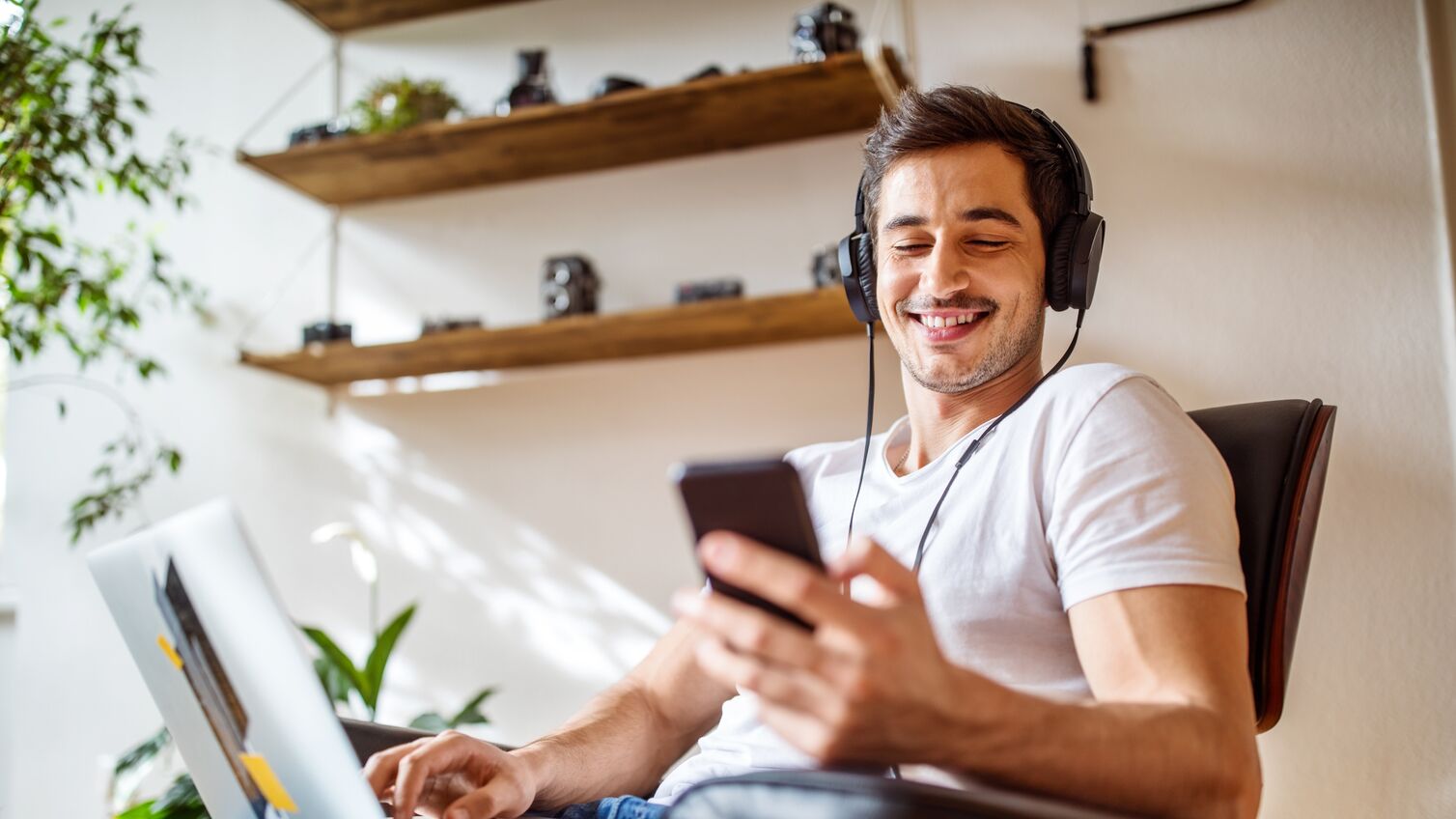 Young man wearing headphones listening to music from mobile phone while working on laptop at home.