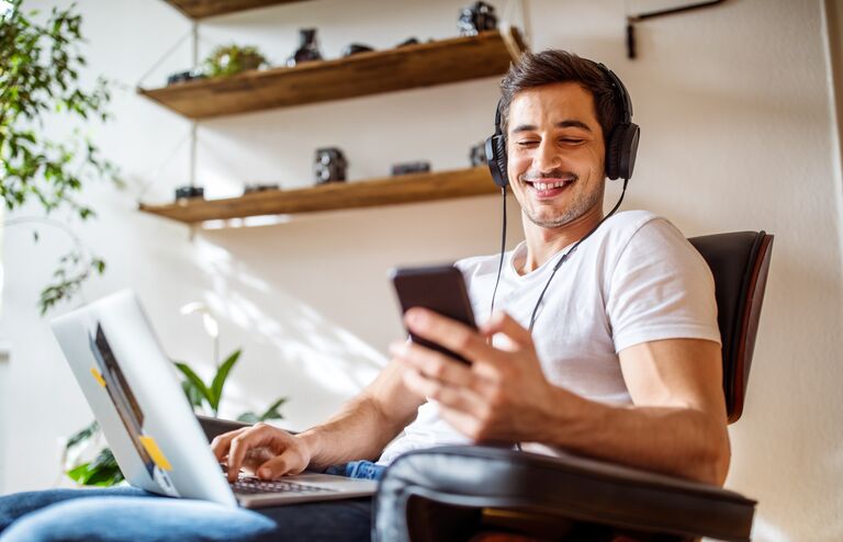 Young man wearing headphones listening to music from mobile phone while working on laptop at home.