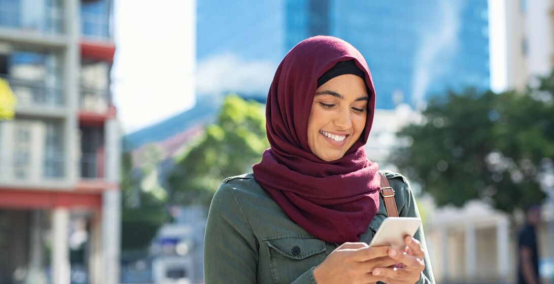 Pretty muslim woman using mobile phone outdoor. Arabic woman wearing hijab using smartphone on the street. Islamic girl texting a message phone in city centre.