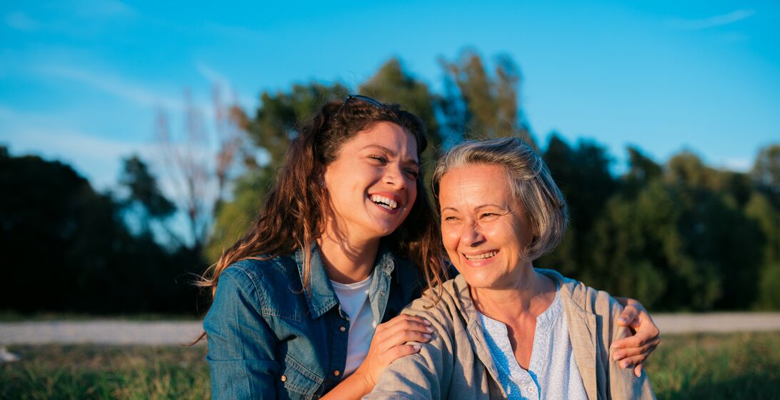 Portrait of beautiful young woman and mother sitting outside in nature. Loving mother and daughter smiling on grass lawn outdoors.