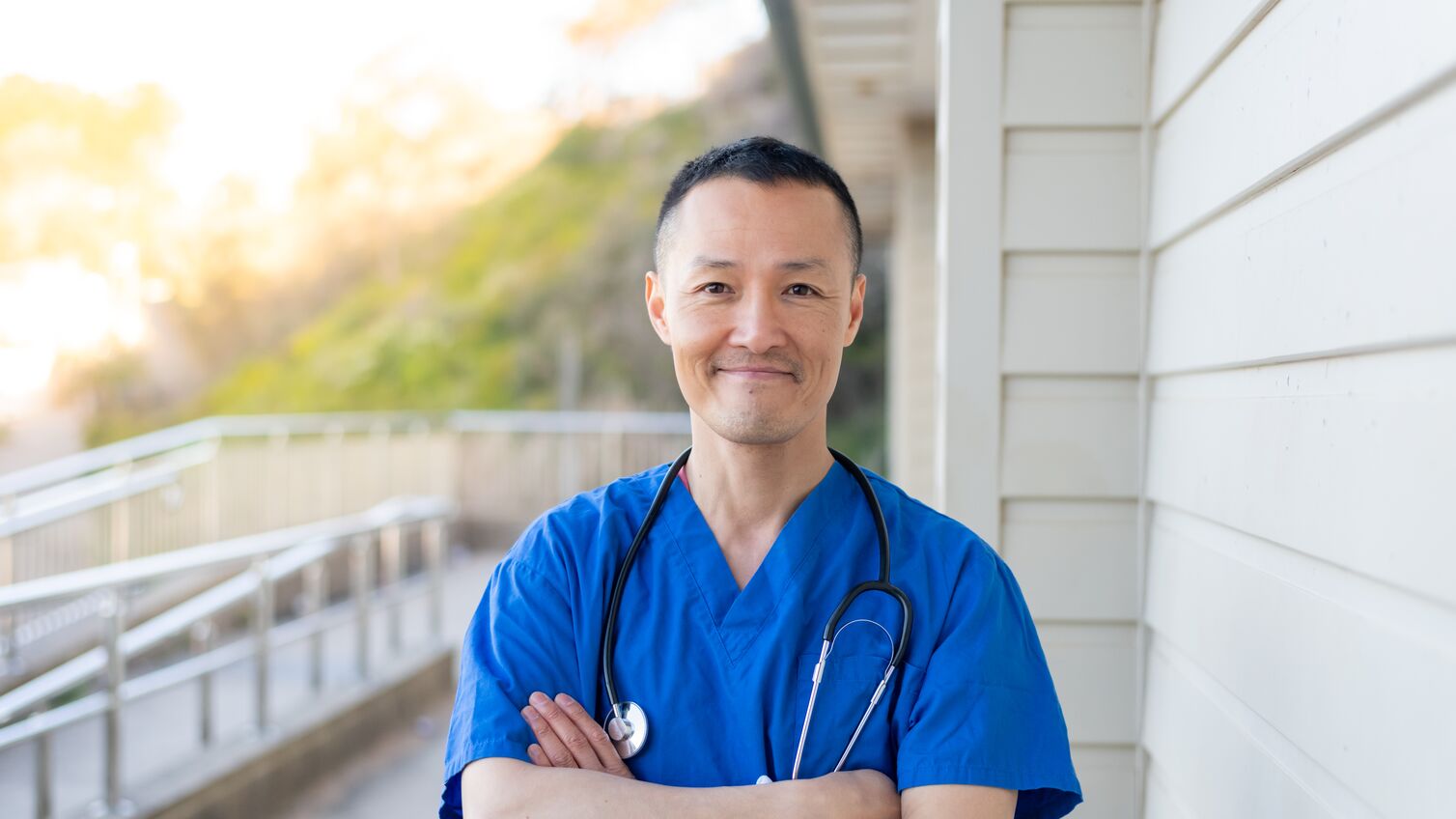 Portrait of a GP, looking directly at the camera and smiling. Standing in front of a hospital.