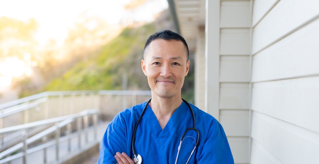 Portrait of a GP, looking directly at the camera and smiling. Standing in front of a hospital.