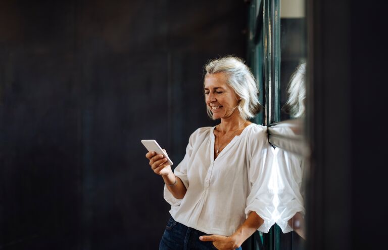 Smiling casual mature businesswoman using smartphone at the window in loft office