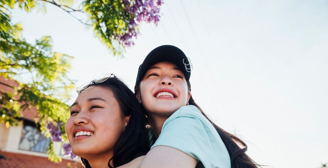 Teenage girls hanging out together walking along a neighbourhood street. One girl is giving the other a piggyback as they laugh together enjoying summer.