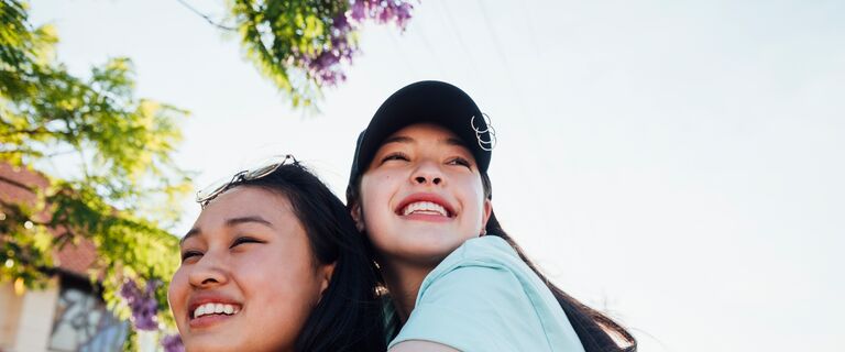 Teenage girls hanging out together walking along a neighbourhood street. One girl is giving the other a piggyback as they laugh together enjoying summer.
