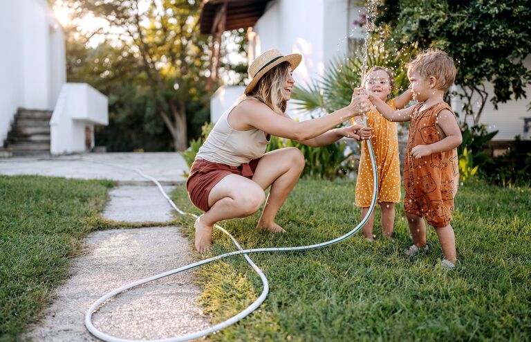 Mother and her cute little children playing with water with garden hose in a backyard