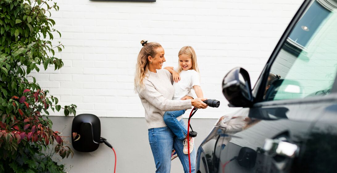 Happy woman carrying girl while walking towards electric car with charger