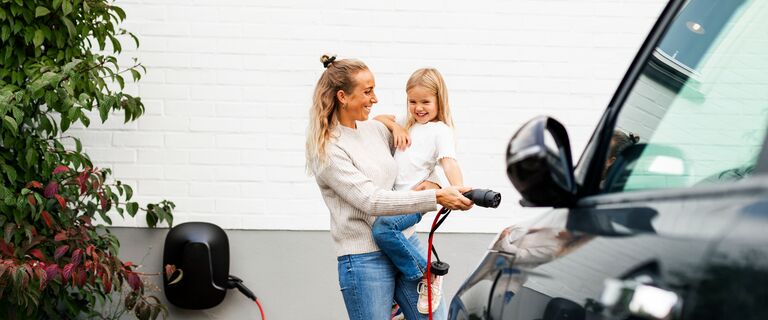 Happy woman carrying girl while walking towards electric car with charger