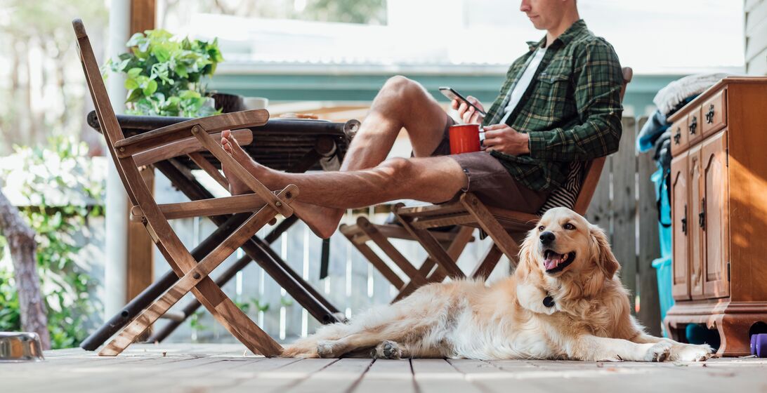 A side view shot of a young man sitting down on a chair outside his house using his smart phone with his dog.