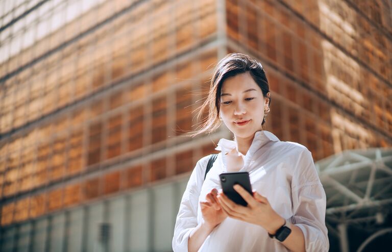 Low angle portrait of young Asian businesswoman checking emails on smartphone outside office building in financial district. With contemporary corporate skyscrapers in background. Making business connections throughout the city